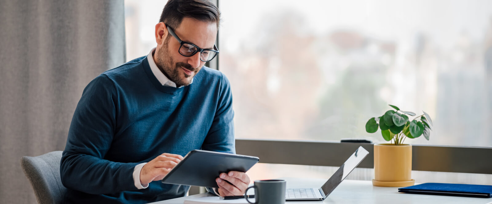 A man holding a tablet and sitting at a table with a laptop on it