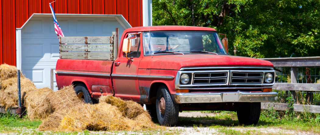 An old red pickup truck with an American flag on it. It sits in front of a red barn.